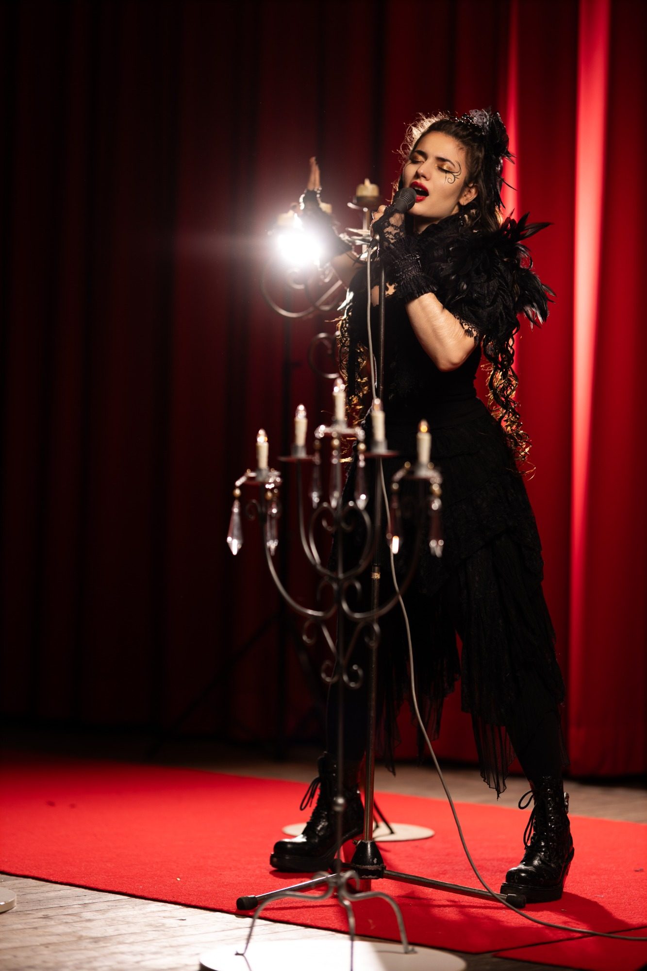 Model: Ella James Frau mit schwarzen Kleid und Federbesatz singt vor einer Kerzenleuchterbühne.